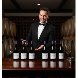 Un sommelier élégant, vêtu d'un smoking noir et d'un nœud papillon, souriant. Il tient un tire-bouchon dans sa main droite et sa main gauche est ouverte en un geste de présentation. Il se tient derrière une table en bois foncé et brillante, sur laquelle sont alignées six bouteilles de vin rouge "Les parcelles". Deux verres à vin vides sont également posés sur la table, ainsi qu'un seau à glace métallique. Une cave à vin, avec des fûts en bois empilés sur les côtés et des casiers à bouteilles en bois remplis de vin au centre. L'éclairage est tamisé, créant une ambiance chaleureuse et sophistiquée, typique d'une dégustation de vin ou d'un environnement de sommellerie.