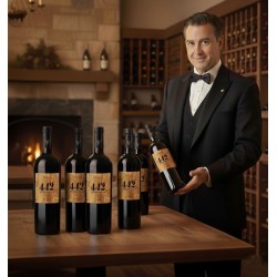 Un sommelier élégamment vêtu d'un smoking noir et d'un nœud papillon, souriant et tenant une bouteille de vin rouge dans une cave à vin. Sur une table en bois, cinq autres bouteilles de vin rouge. Toutes les bouteilles portent une étiquette distinctive avec le numéro "442 PECH NAUT".On aperçoit une cheminée avec un feu allumé, ajoutant une ambiance chaleureuse, ainsi que des casiers à vin remplis de bouteilles, suggérant une collection étendue. L'éclairage est doux, mettant en valeur les bouteilles et le sommelier, et créant une atmosphère sophistiquée et accueillante.