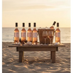 Plage de sable fin, au coucher du soleil. Une table en bois est installée directement sur le sable.Sur cette table, six bouteilles de vin rosé syrah sont joliment disposées. Un panier en osier tressé, rempli de glace, sert de seau pour rafraîchir l'une des bouteilles. Deux verres à vin remplis de rosé.Coquillages et brins de lavande sur la table La mer.
