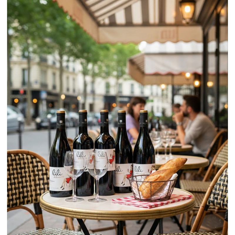 Sur la table ronde d'un café parisien, six bouteilles de vin rouge "Lili Bio" sont mises en avant, accompagnées de deux verres vides et d'une corbeille contenant une baguette fraîche. La scène se déroule sur une terrasse typique, avec ses chaises en rotin et son auvent rayé, donnant sur une rue animée bordée d'arbres et d'immeubles haussmanniens.