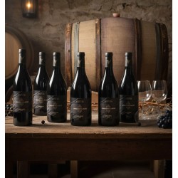 six bouteilles L'enclos des Lauzes Cabardès cave a vin Vendéole élevé en fût de chêne disposées sur une table en bois rustique.Des murs en pierre et de grands fûts de chêne, créant une atmosphère authentique et traditionnelle.On trouve des verres à vin vides, dont un est renversé, et des grappes de raisin noir, suggérant une dégustation récente ou à venir. L'éclairage est tamisé, avec une source de lumière chaude provenant d'une lanterne en arrière-plan, ce qui ajoute une ambiance chaleureuse et intime à la composition.