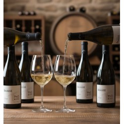 Dégustation de vin dans une cave ou un chai.Six bouteilles, Deux grands verres à vin sur pied sont en train d'être remplis simultanément de vin blanc chardonnay par deux bouteilles distinctes. Les bouteilles, de couleur sombre, ont une étiquette blanche "CHÂTEAU BRASSE" et une illustration de ce qui ressemble à un domaine viticole. D'autres bouteilles de vin rangées sur des étagères et un grand tonneau en bois. La table sur laquelle reposent les verres est en bois rustique. L'éclairage est chaleureux et met en valeur les couleurs dorées du vin.