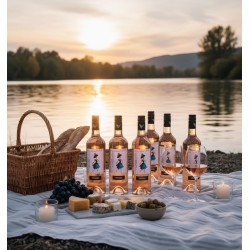 Pique-nique idyllique, au bord d'un lac, baignée par la lumière chaude et enveloppante d'un coucher de soleil. Une couverture blanche à carreaux est étendue sur un sol caillouteux ou sablonneux. Six bouteilles de vin rosé, révèlent un design distinctif orné d'une illustration stylisée d'une femme et le nom "VENDÉOLE" deux verres à pied, partiellement remplis et prêts à être dégustés. L'ensemble est complété par un panier en osier tressé, contenant deux baguettes de pain, et une planche en marbre présentant un assortiment de fromages variés. Une grappe de raisins noirs et un petit bol d'olives vertes ajoutent des touches de couleur et de fraîcheur à cette composition gourmande. Deux petites bougies blanches, placées dans des photophores en verre, diffusent une lumière douce et chaleureuse, renforçant l'ambiance intime et romantique.